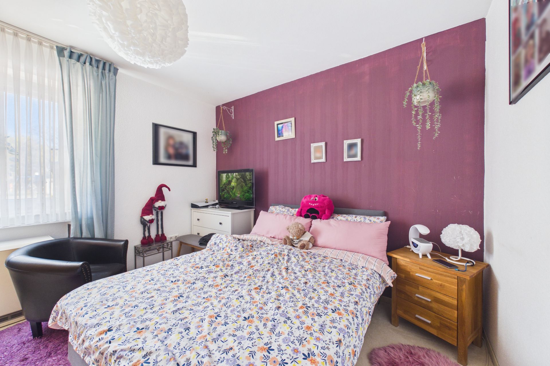 Schlafzimmer mit Doppelbett, lilafarbener Wand, Nachttisch, Sessel und Dekoration.