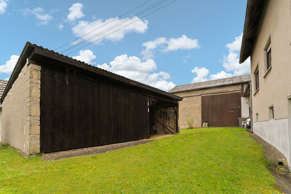 Hof mit Rasenfläche, Holzhütte und Nebengebäude neben einem Wohnhaus bei blauem Himmel.
