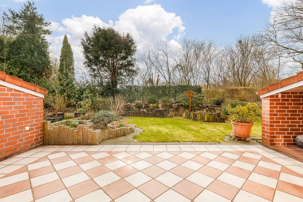 Blick von einer gefliesten Terrasse auf einen Garten mit Rasen, Beeten und Bäumen.