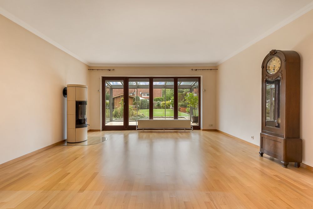 Helles Wohnzimmer mit Holzfußboden, Kaminofen, Standuhr und großen Fenstern zum Garten.