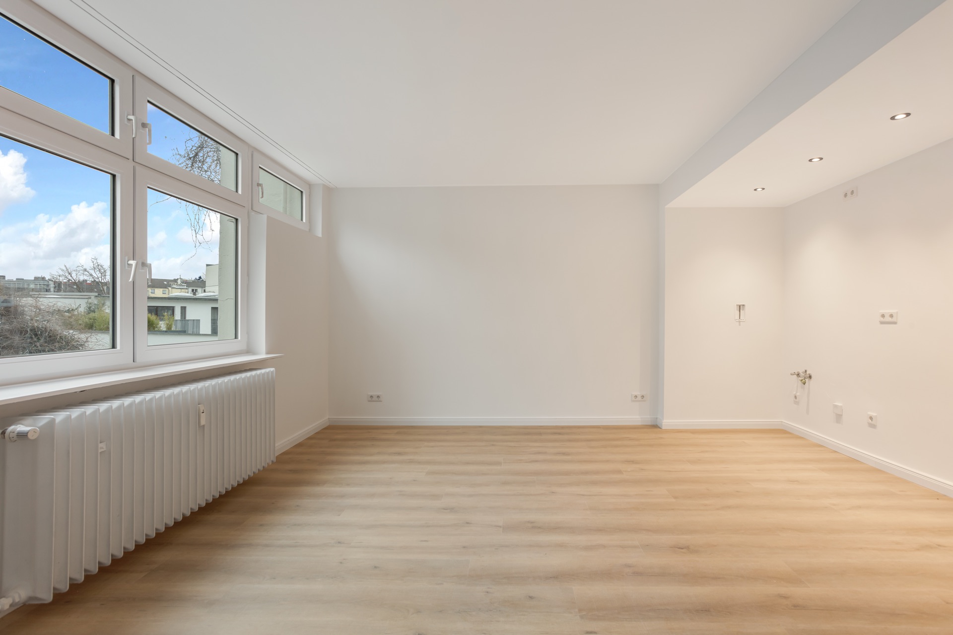 Helles, leeres Zimmer mit großen Fenstern, Parkettboden und Heizkörper.