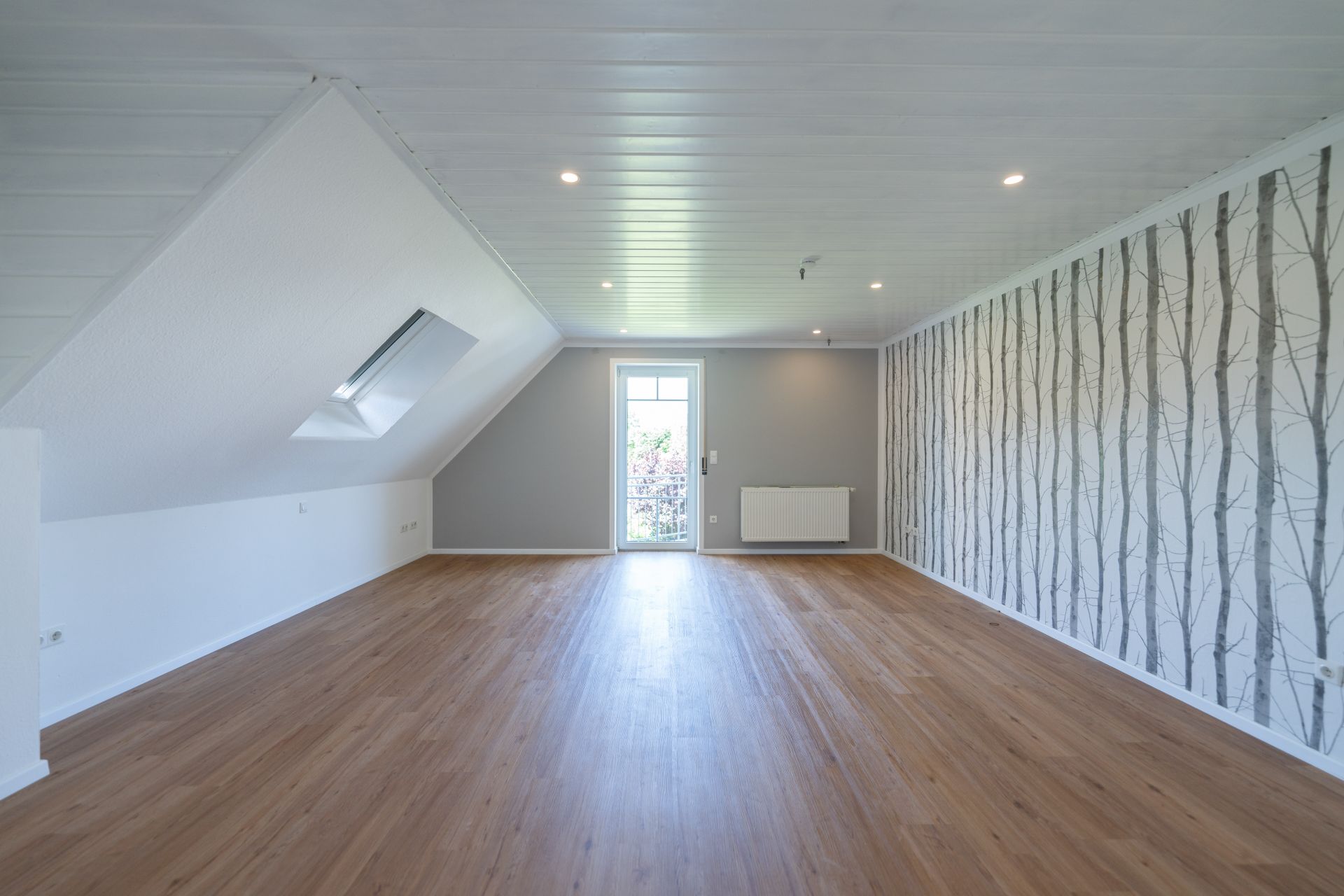 Heller, leerer Dachgeschossraum mit Holzfußboden und gemusterter Tapete an einer Wand.