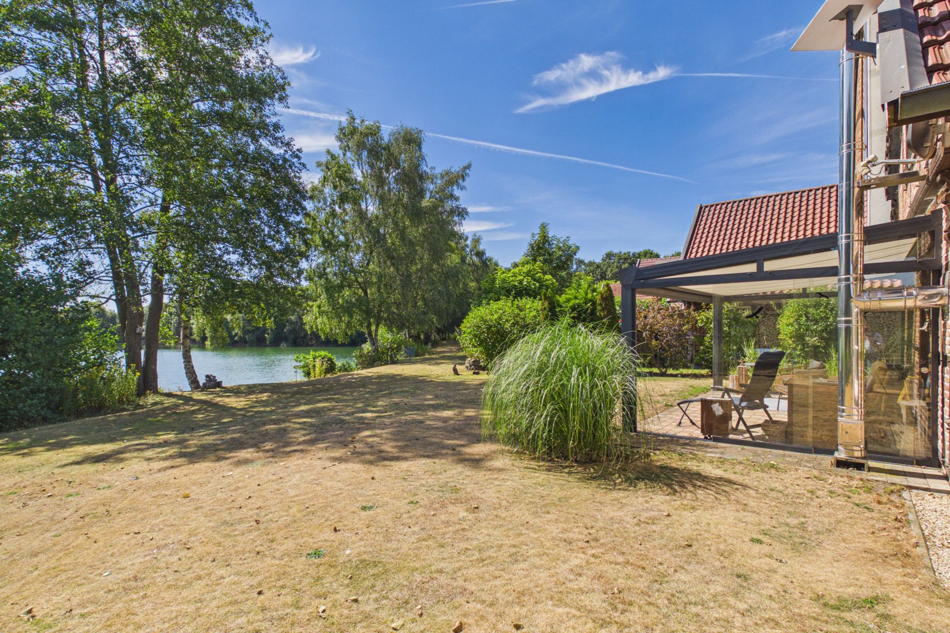 Garten mit Rasenfläche, Bäumen, Wintergarten und Blick auf einen See an einem sonnigen Tag.