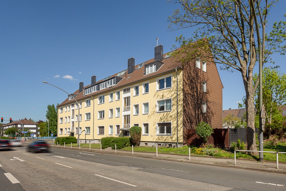 Mehrfamilienhaus mit gelber Fassade an einer Straße, Bäume und Autos im Vordergrund.