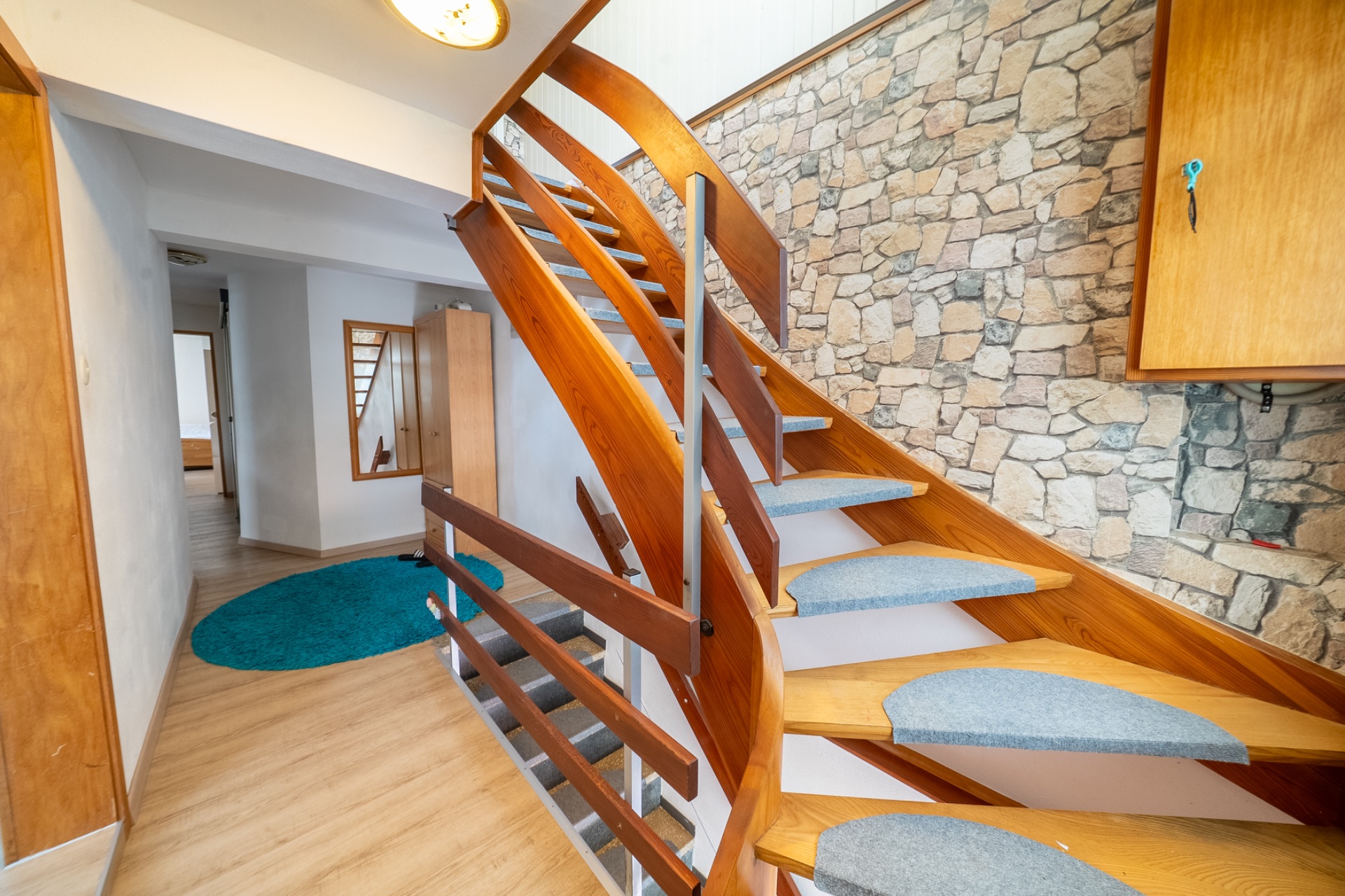 Holztreppe mit grauen Stufenmatten vor einer Steinwand in einem Flur mit Parkettboden.