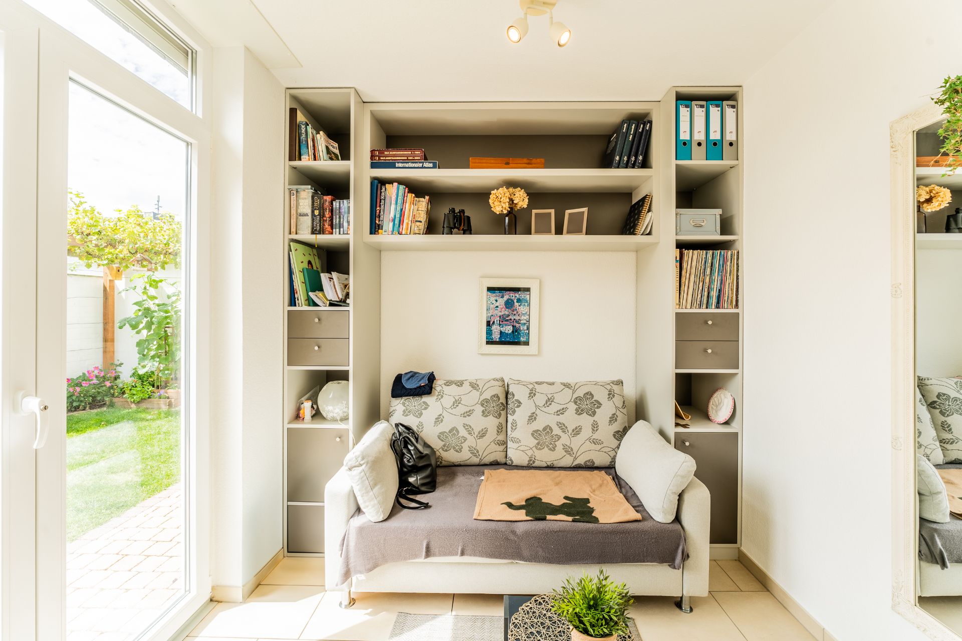 Helles Wohnzimmer mit Sofa, eingebauten Regalen, Glastür und Blick in den Garten.