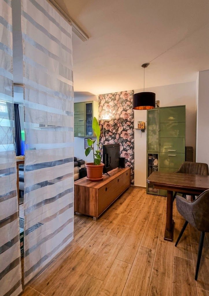 Wohnzimmer mit Holzfußboden, Holzmöbeln, grünem Schrank und floraler Tapete.