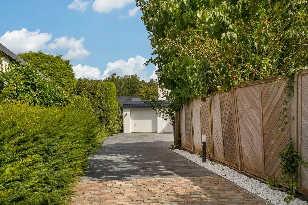 Gepflasterte Einfahrt mit Holzzaun und grüner Bepflanzung, Garage im Hintergrund.
