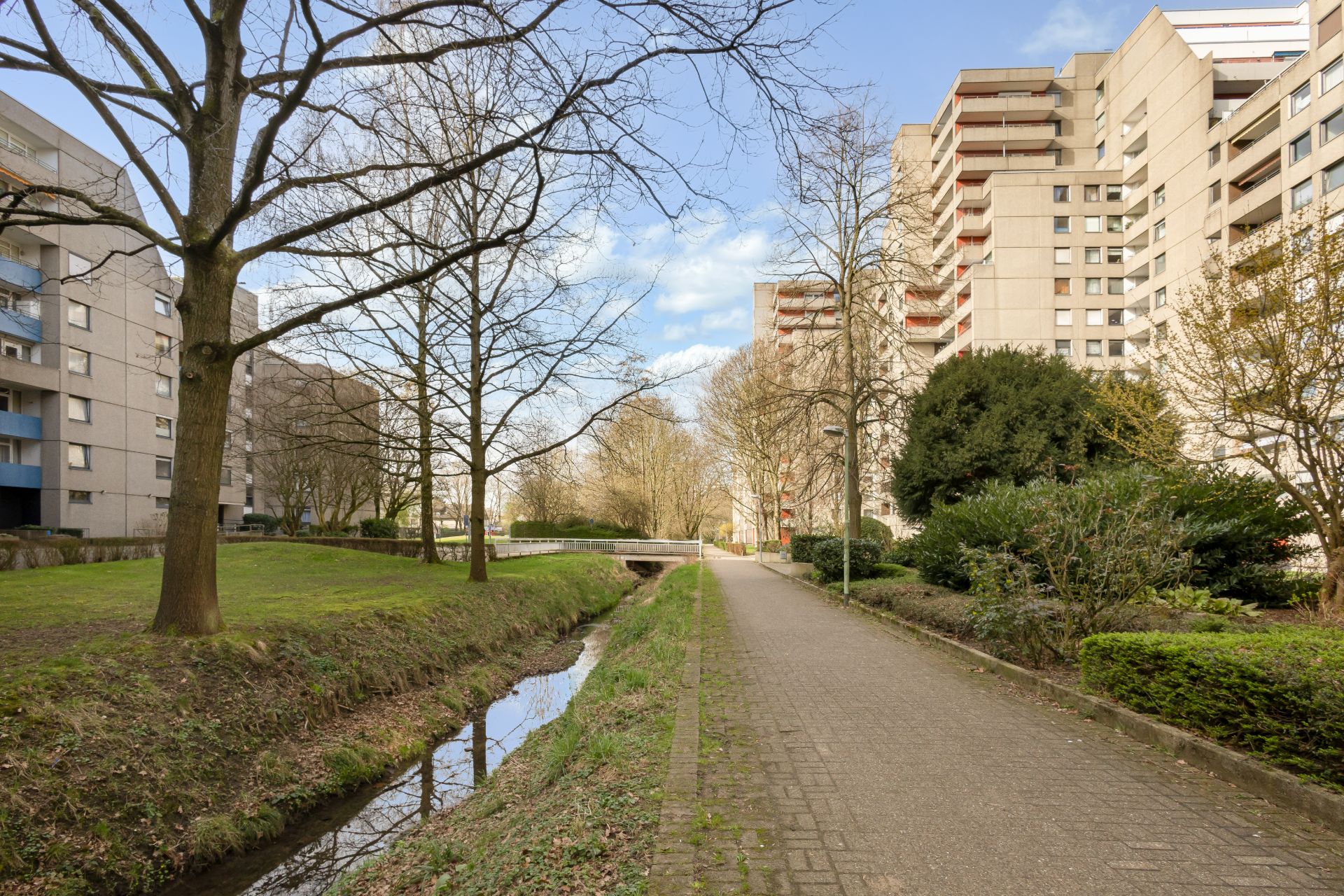 Gepflasterter Weg mit Bach, Bäumen und Wohngebäuden im Hintergrund.