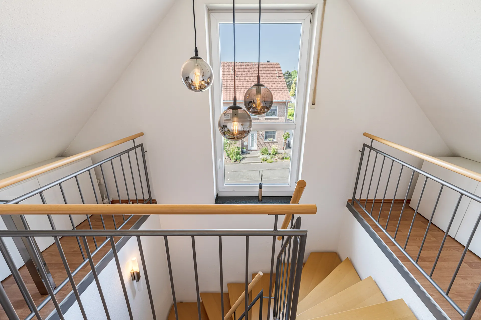Treppenhaus mit Holzstufen, Geländer und großen Fenstern, drei runde Lampen hängen von der Decke.