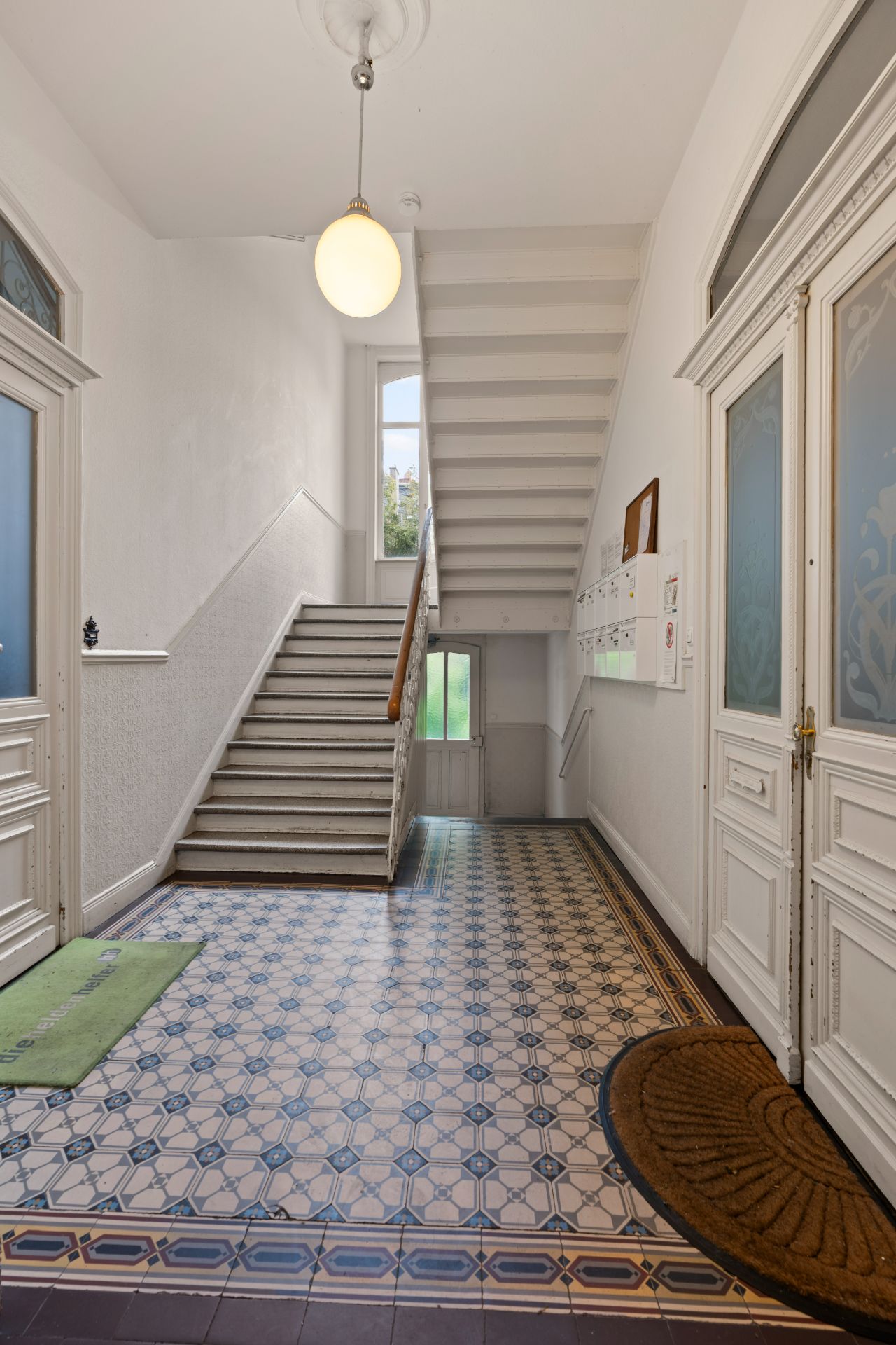 Flur mit gemustertem Fliesenboden, Treppe und weißen Türen in einem Wohnhaus.
