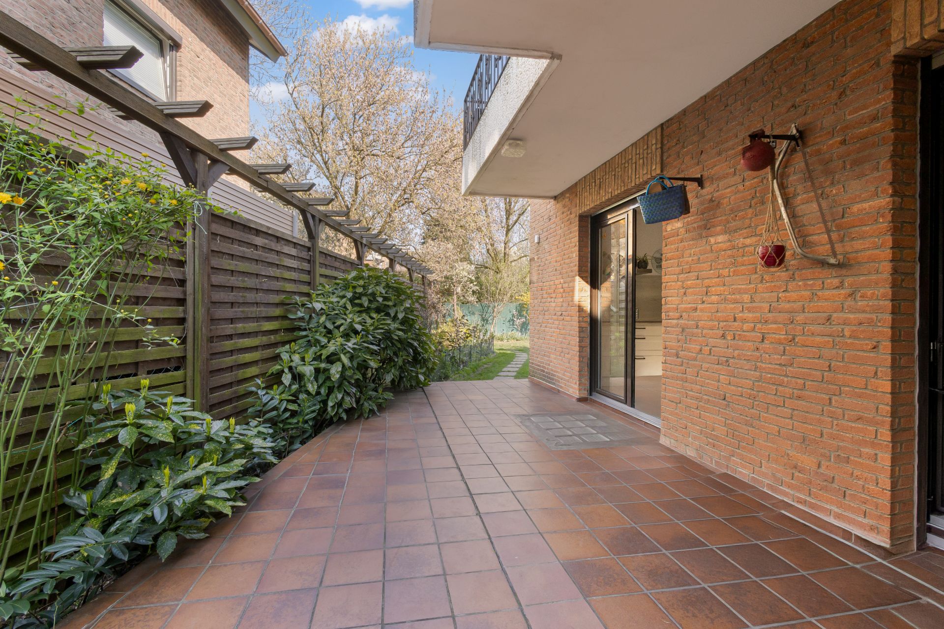 Geflieste Terrasse mit Pflanzen, Holzzaun und Zugang zu einem Garten, angrenzend an ein Backsteinhaus.