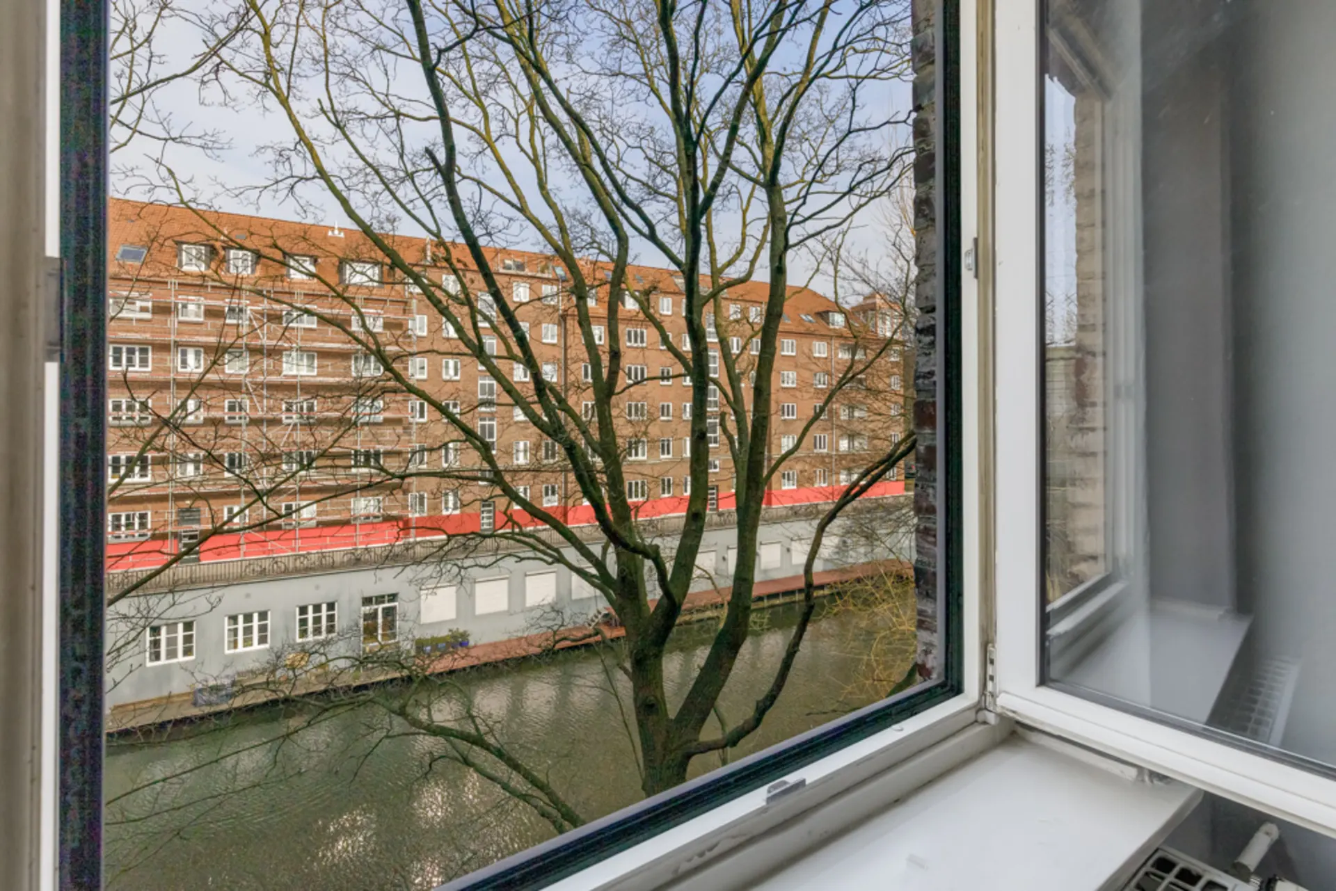 Blick aus offenem Fenster auf einen Kanal, Bäume und ein Mehrfamilienhaus.