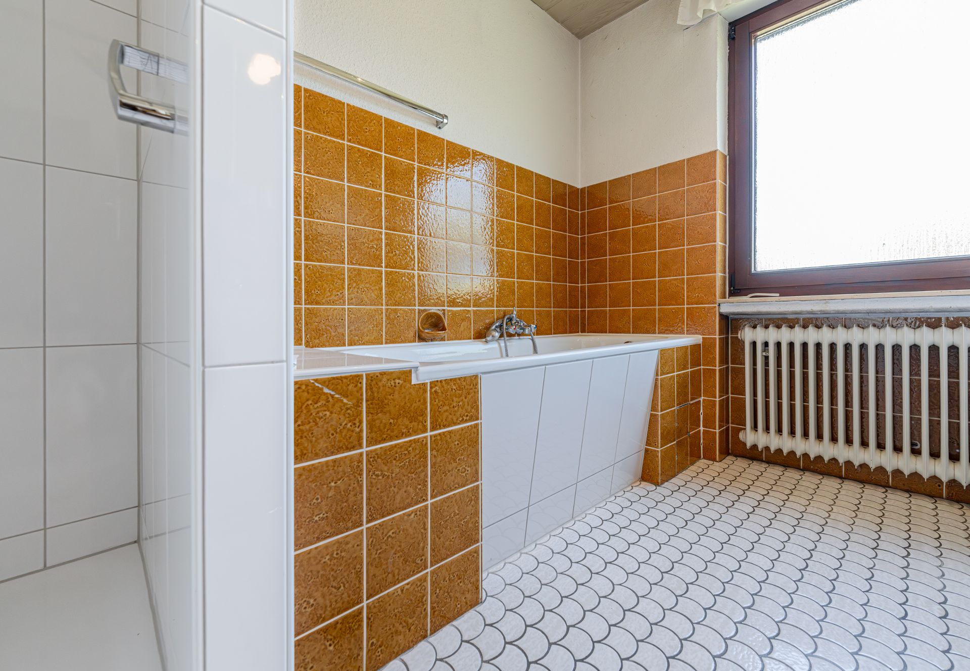 Badezimmer mit Badewanne, braunen Fliesen und Fenster mit Heizung darunter.