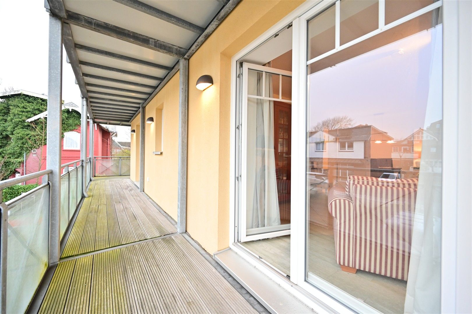 Überdachter Balkon mit Holzfußboden und Geländer, Zugang durch eine große Glastür, Sessel im Raum sichtbar.