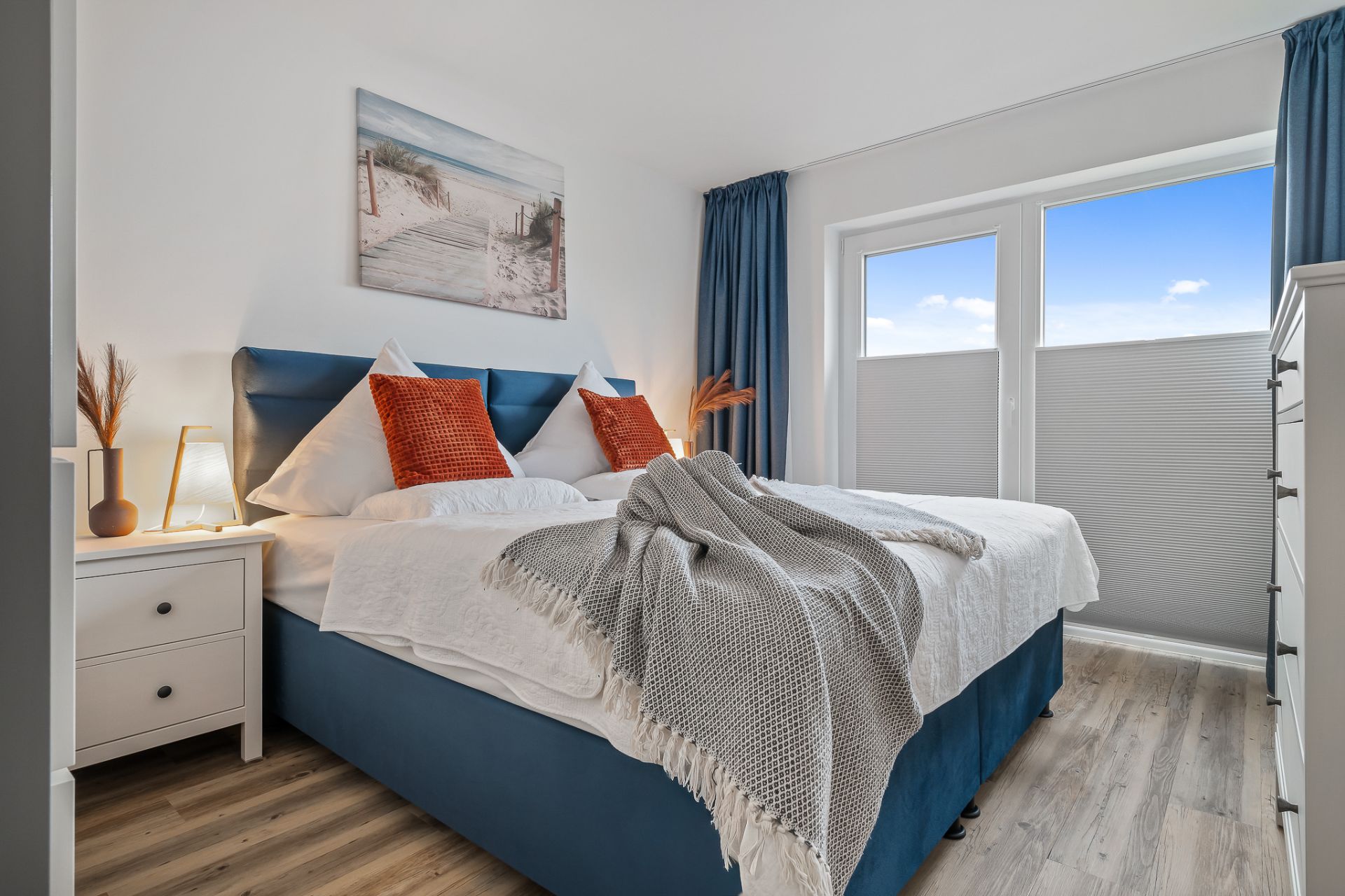Schlafzimmer mit Doppelbett, blauer Bettwäsche, Nachttisch und großem Fenster.