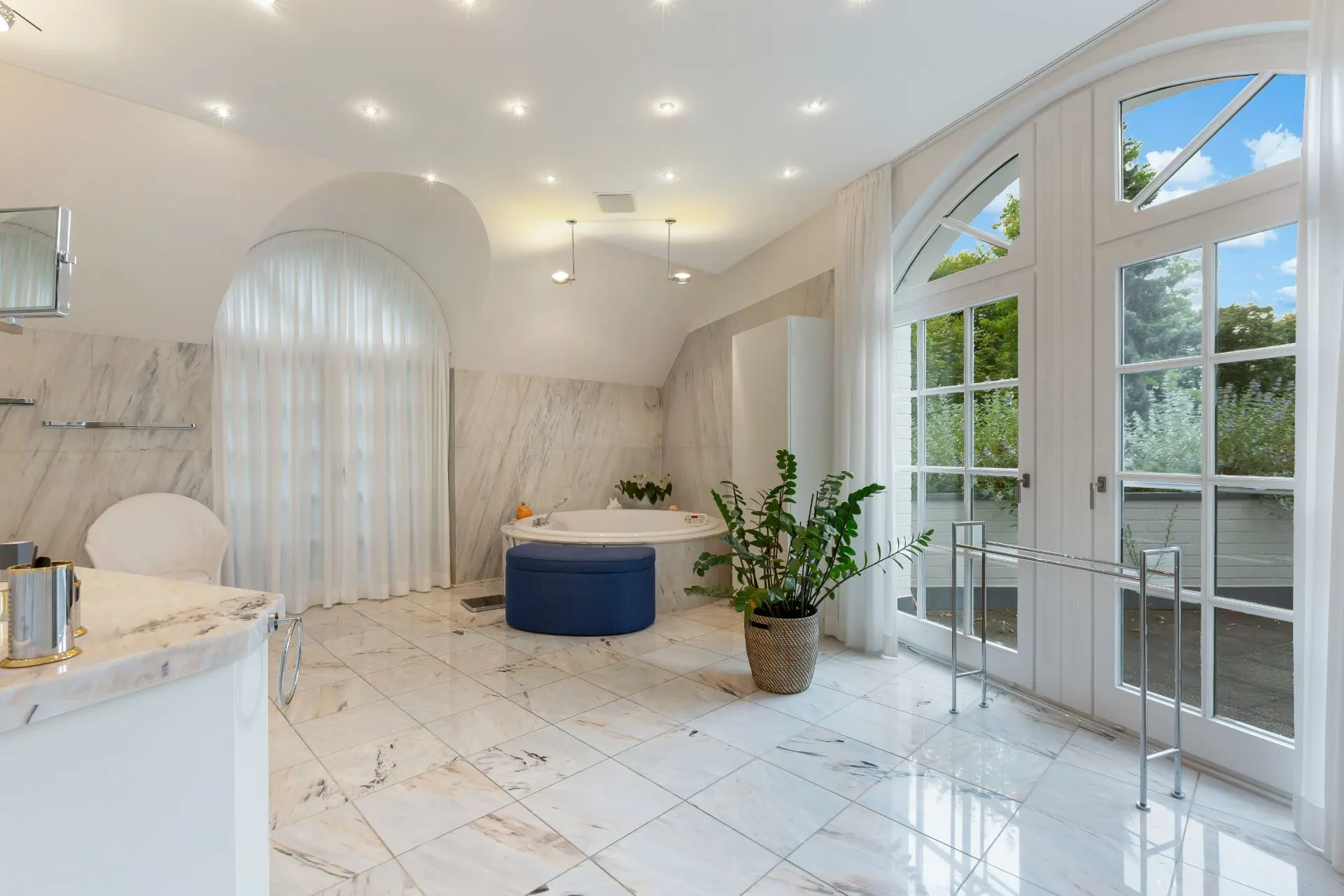 Helles Badezimmer mit Badewanne, großen Fenstern, Marmorfliesen und einer Zimmerpflanze.