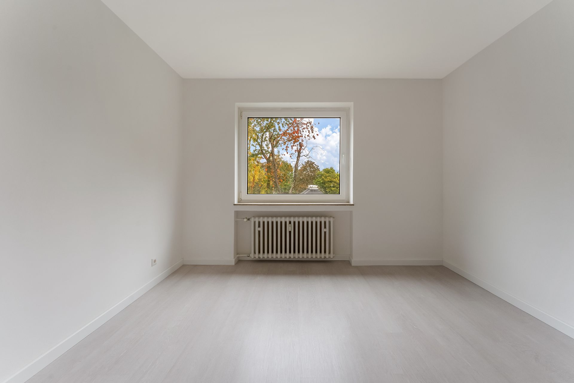 Leerer, heller Raum mit großem Fenster und Heizkörper darunter, heller Boden.