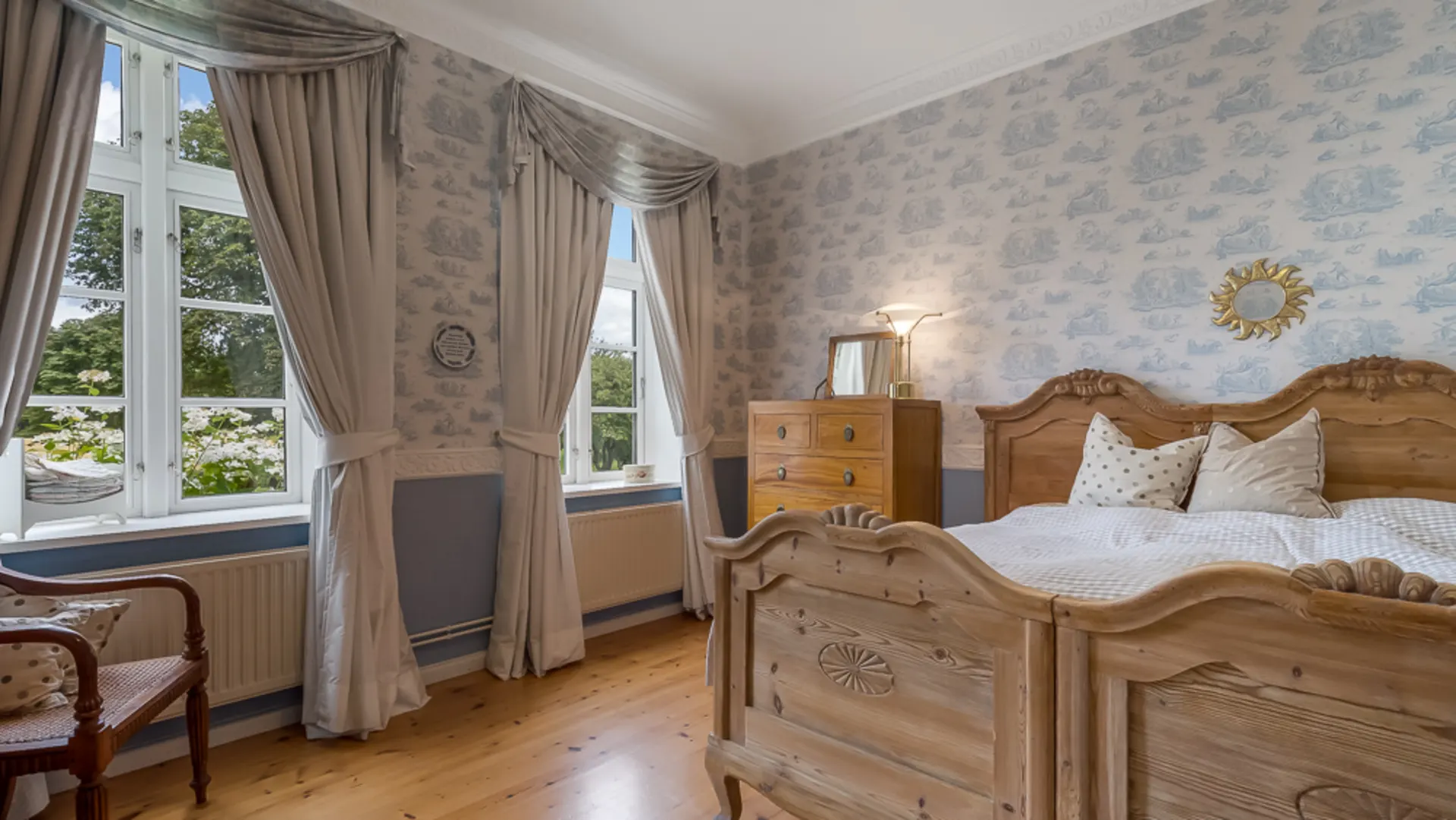 Helles Schlafzimmer mit zwei Holzbetten, Kommode, Sessel und großen Fenstern mit Vorhängen.