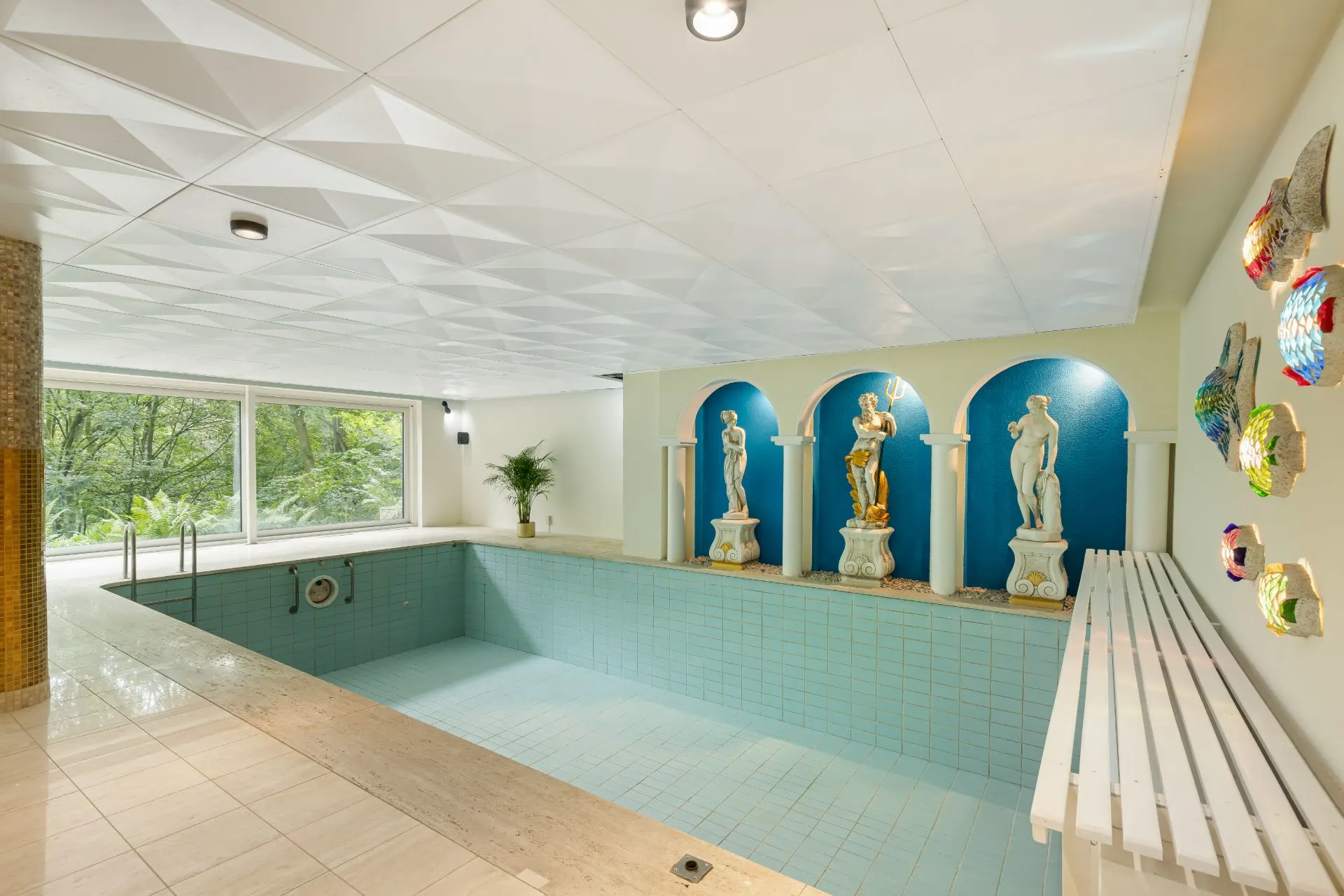 Leeres Hallenbad mit blauen Fliesen, großen Fenstern und vier Statuen an einer Wand.
