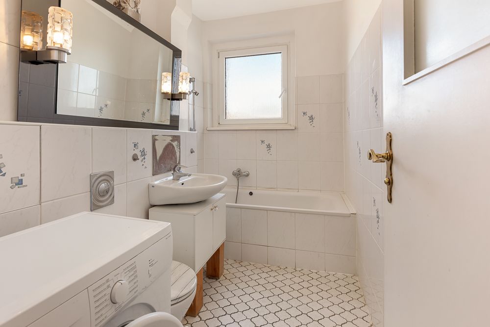 Helles Badezimmer mit Waschmaschine, Waschbecken, Spiegel, Badewanne und Fenster.