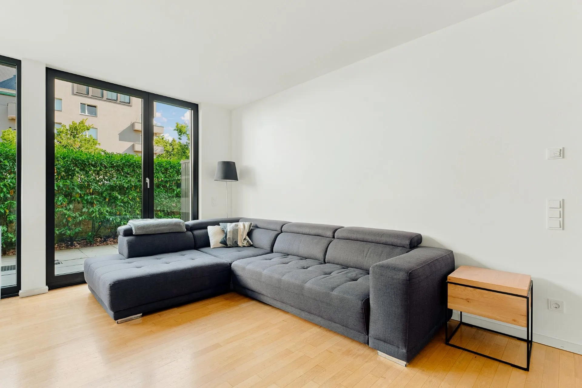 Wohnzimmer mit großer grauer Eckcouch, kleinem Holztisch und bodentiefen Fenstern mit Blick ins Grüne.