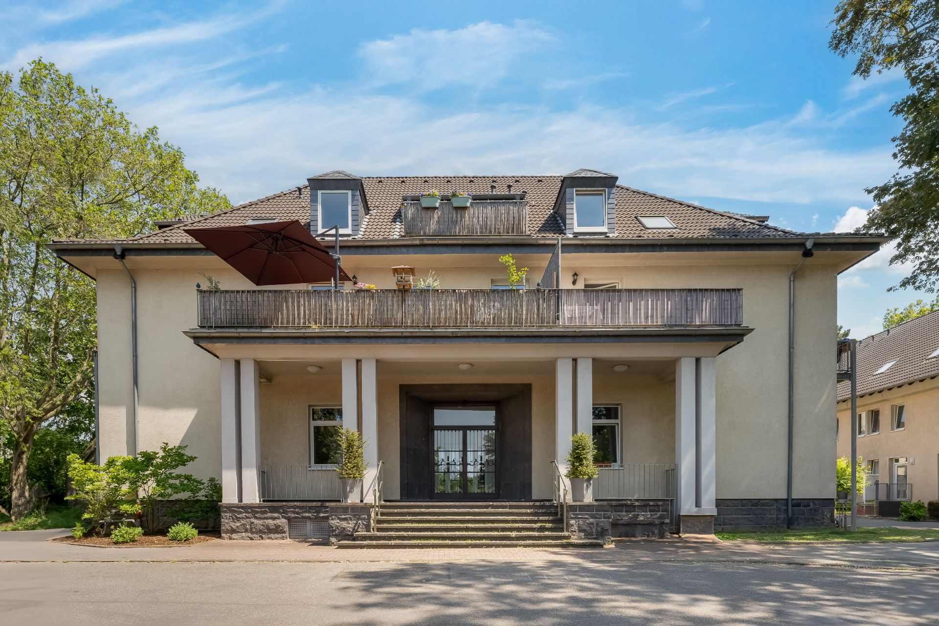 Vorderansicht eines zweistöckigen Wohnhauses mit Balkon, Pflanzen und großem Sonnenschirm.