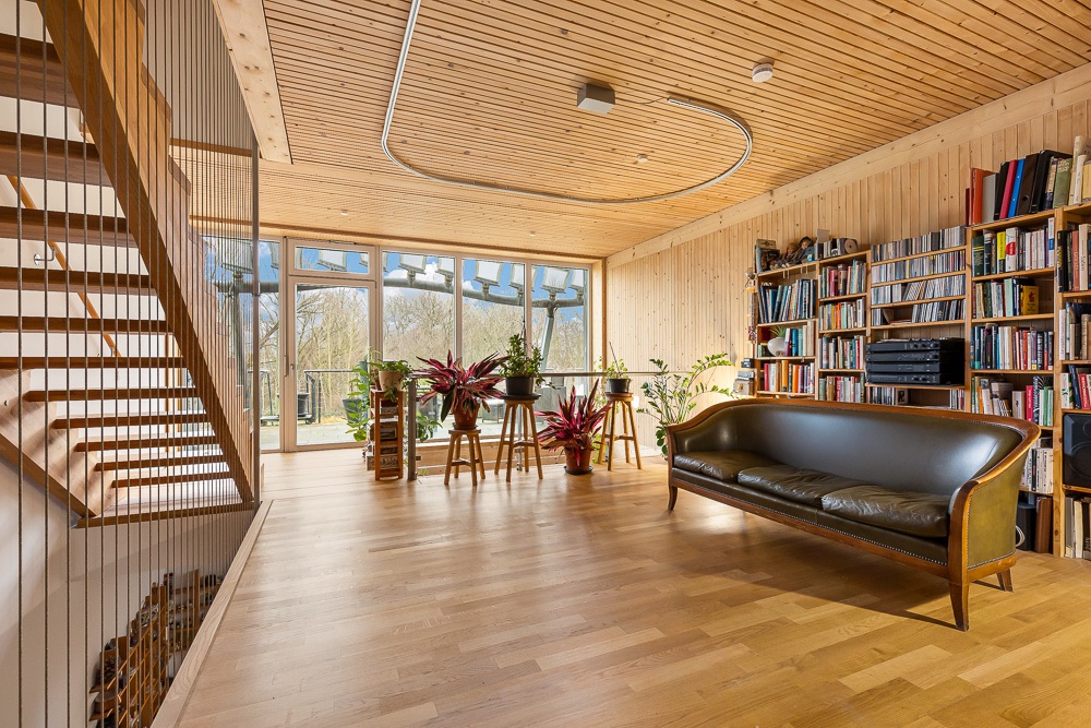 Helles Wohnzimmer mit Holzdecke, Bücherregalen, Sofa, vielen Pflanzen und großer Fensterfront.