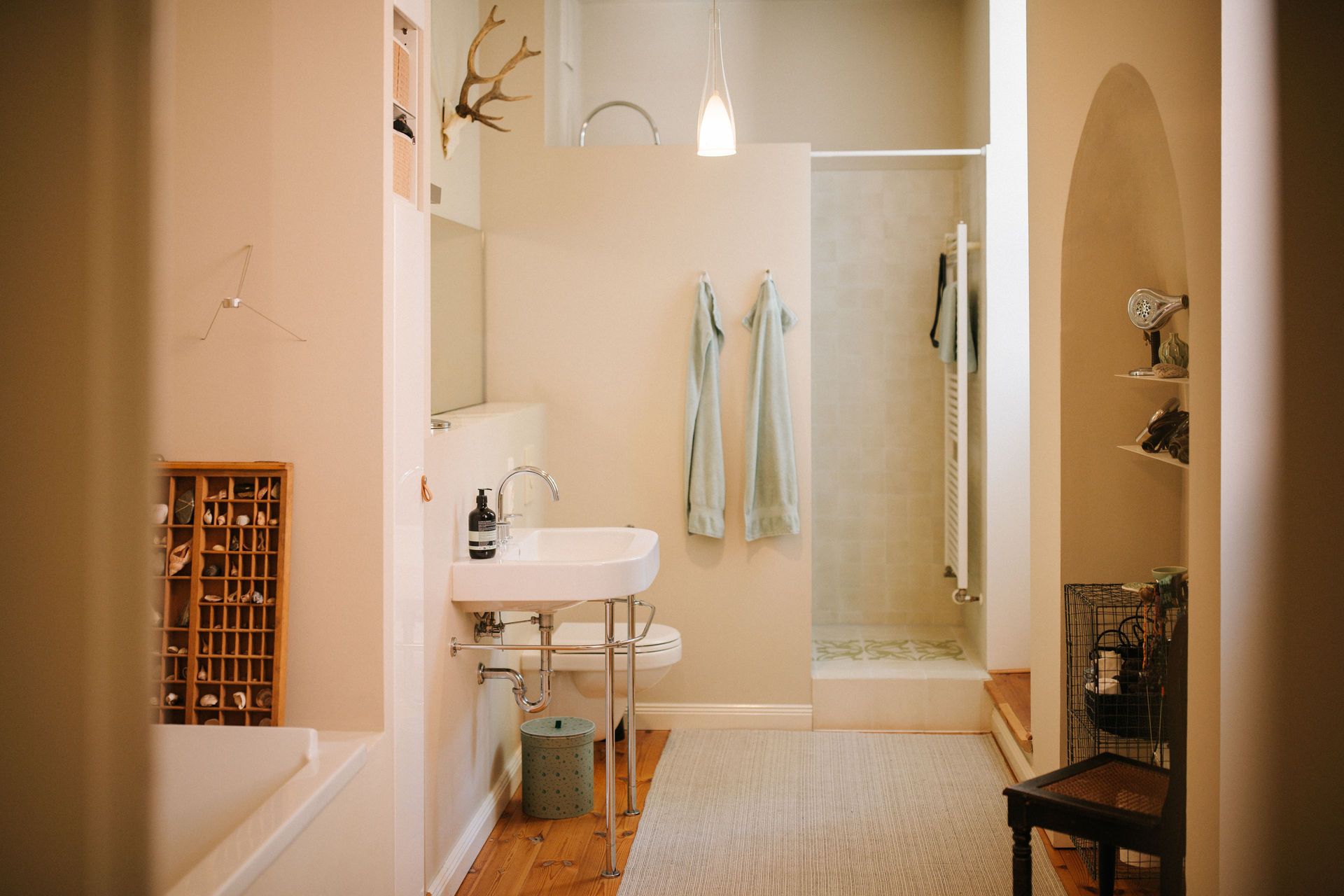 Helles Badezimmer mit Waschbecken, Dusche, Handtüchern und Regalen an der Wand.