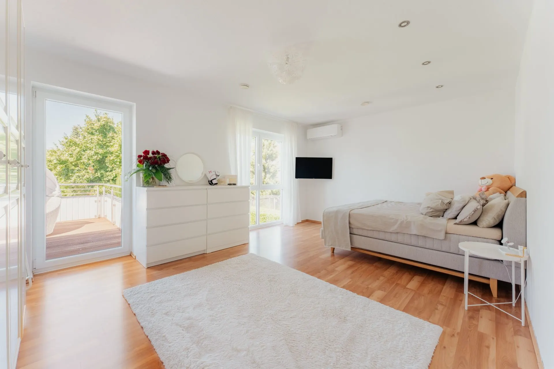 Helles Schlafzimmer mit Bett, Kommode, Wandfernseher und großem Teppich auf Holzboden.