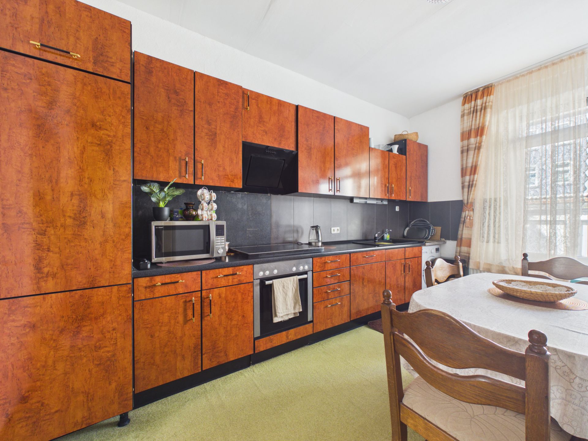 Küche mit braunen Holzmöbeln, Esstisch, Mikrowelle und großem Fenster mit Vorhängen.