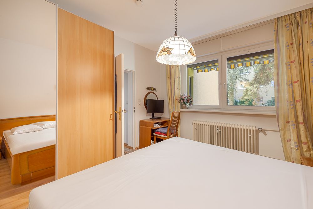 Schlafzimmer mit Doppelbett, Schrank, Schreibtisch und großem Fenster mit Vorhängen.