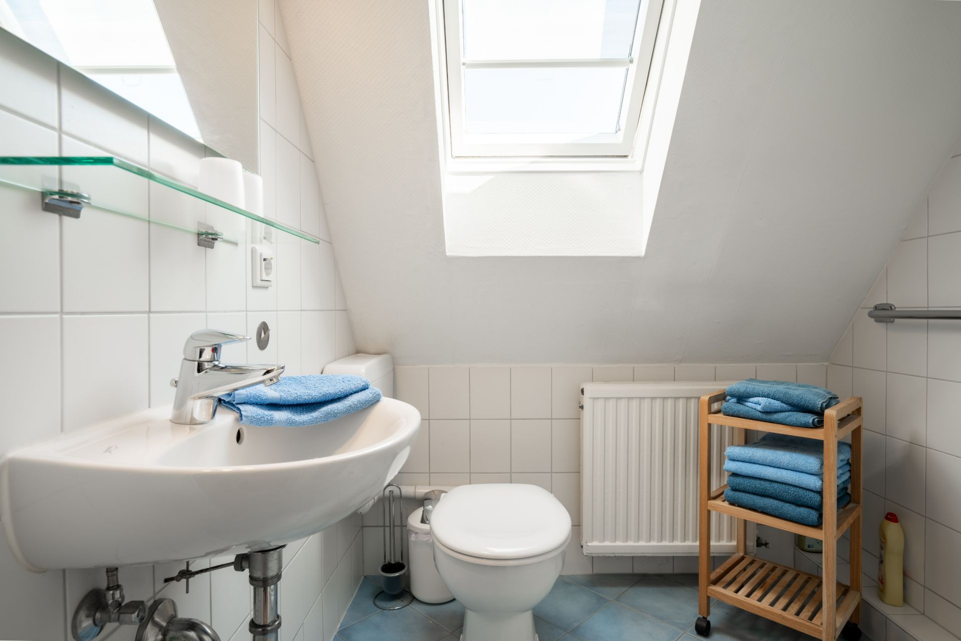 Kleines Bad mit Waschbecken, Toilette, Dachfenster und Regal mit blauen Handtüchern.