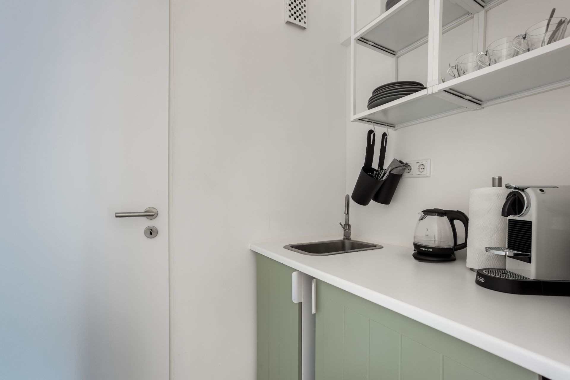Kleine Küchenzeile mit Spüle, Kaffeemaschine, Wasserkocher und offenem Regal mit Geschirr.