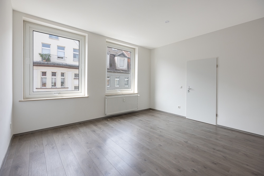 Leerer Raum mit zwei großen Fenstern, weißer Wand und grauem Holzboden.