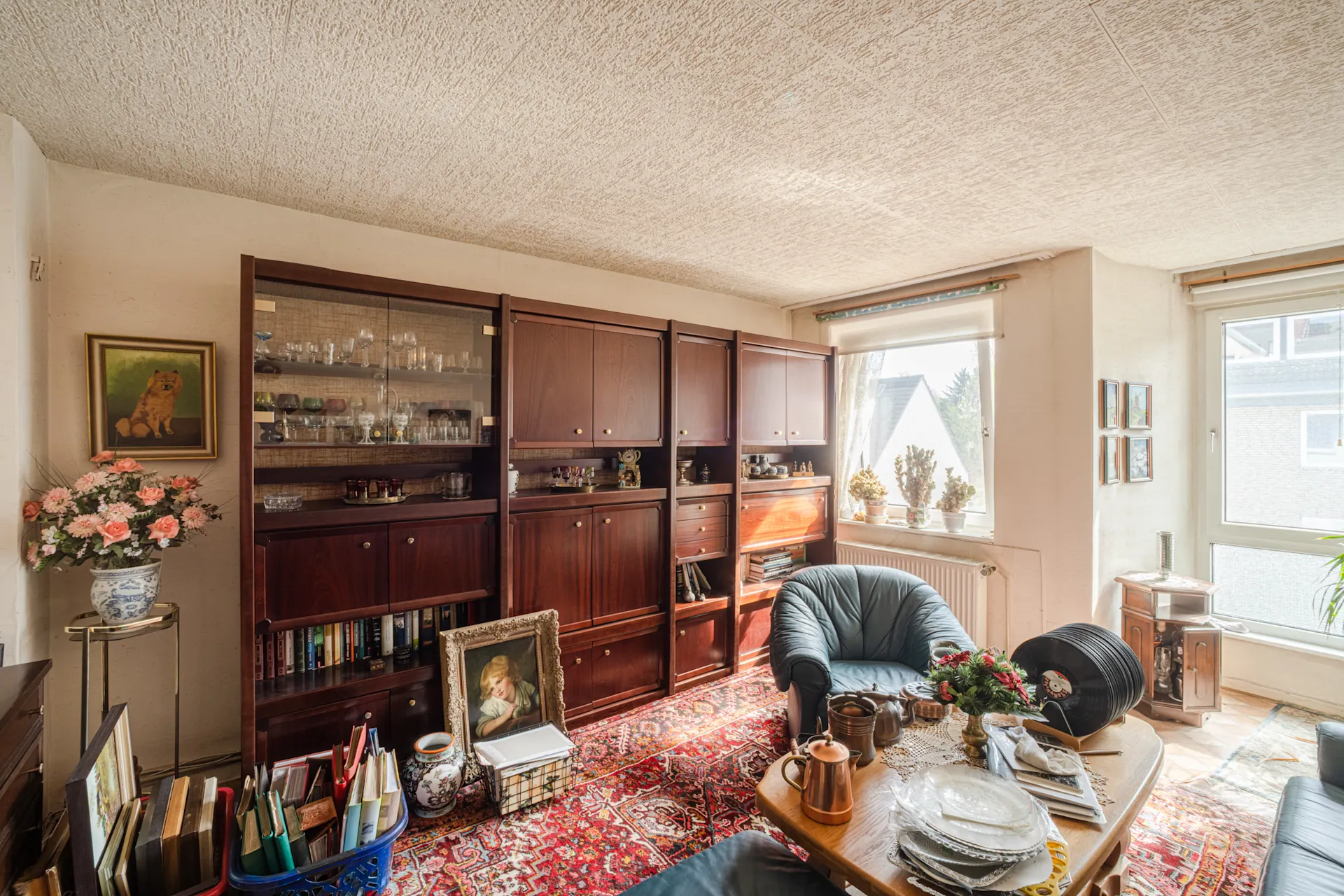 Wohnzimmer mit Teppich, dunklen Regalen, Sessel, Bildern und vielen Büchern bei Tageslicht.