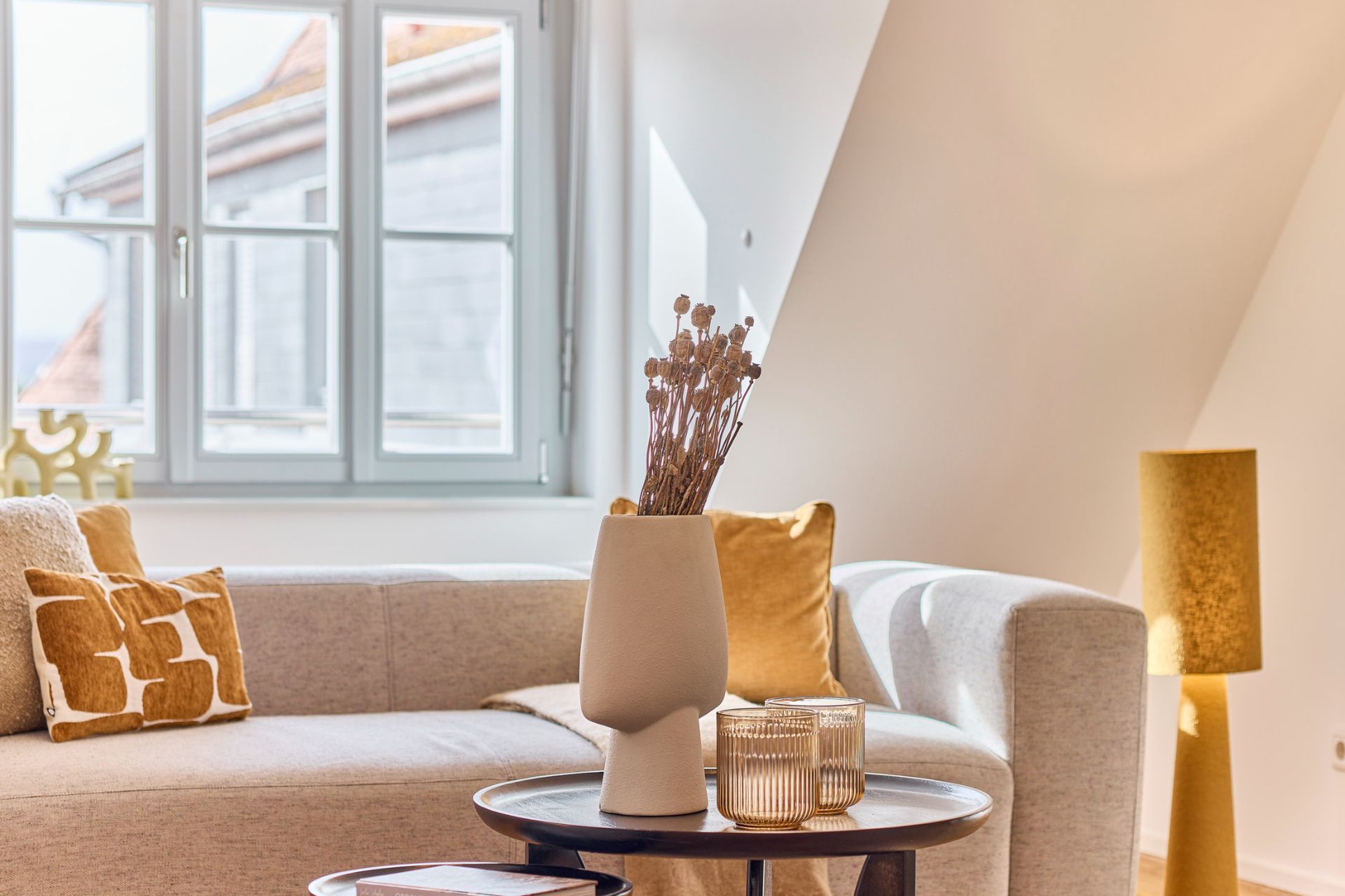 Helles Wohnzimmer mit Sofa, Kissen, Beistelltischen, Vase und Stehlampe vor einem Fenster.