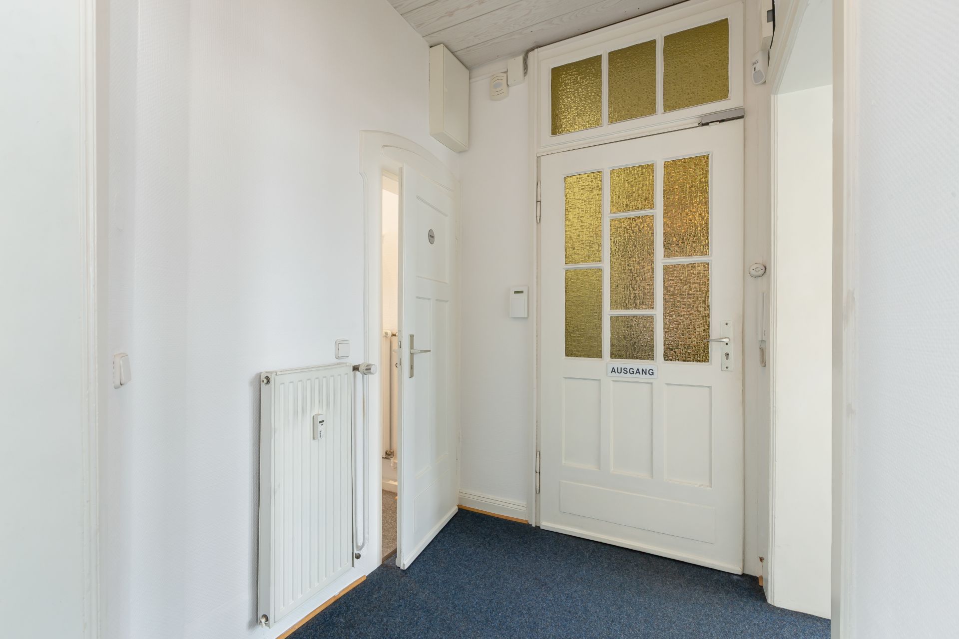 Heller Flur mit weißer Tür, Heizkörper und blauem Teppichboden.