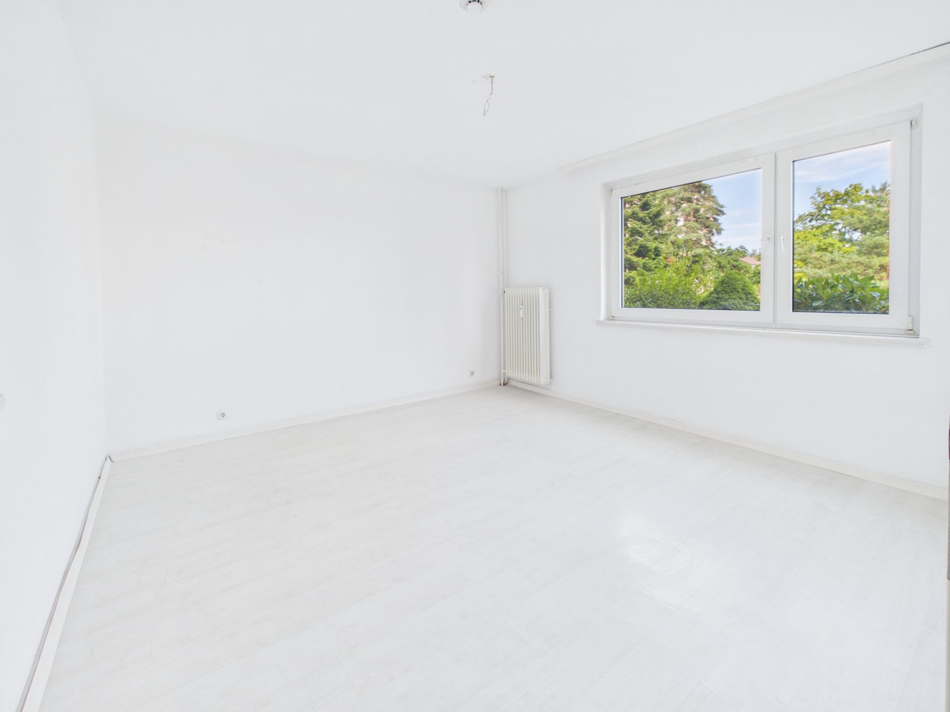 Helles, leeres Zimmer mit weißen Wänden, großem Fenster und Blick auf Bäume.