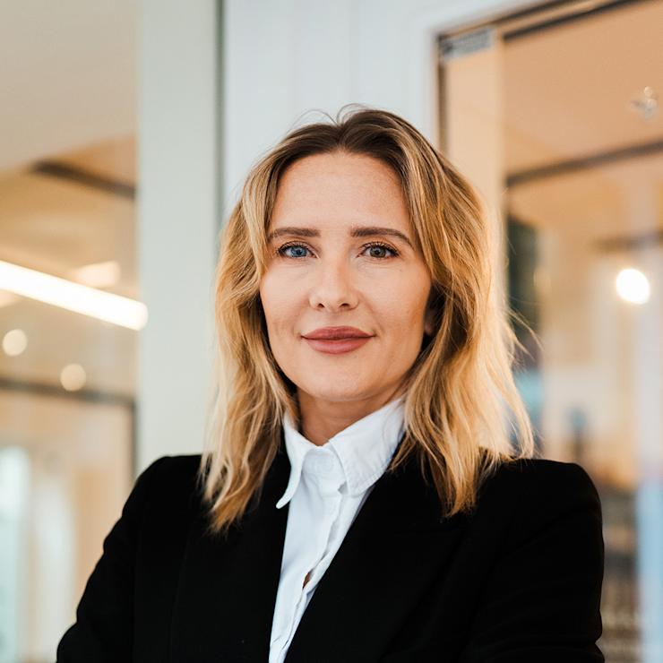 Frau mit blondem Haar in schwarzem Blazer steht mit verschränkten Armen in modernem Innenraum.