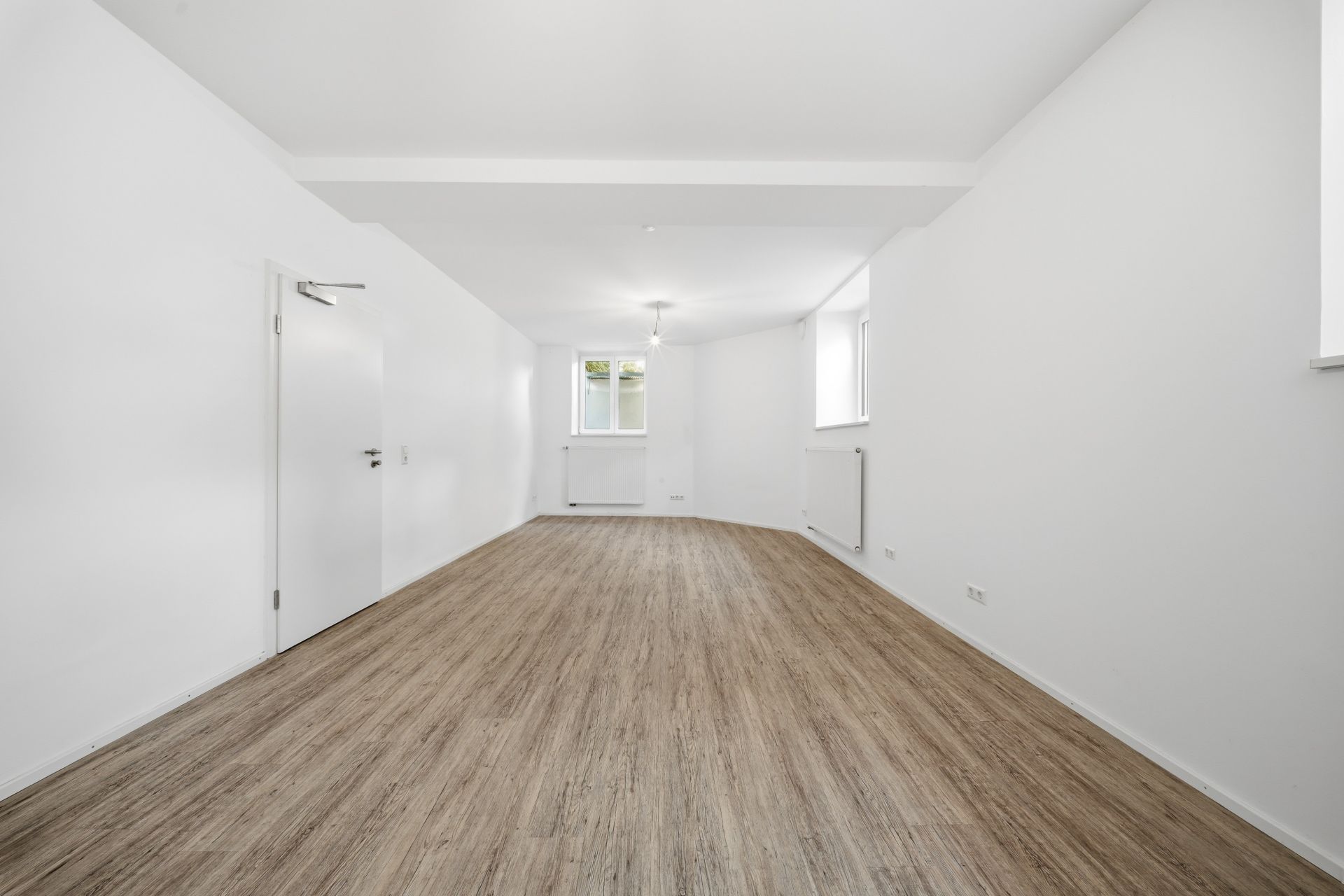 Helles, leeres Zimmer mit Holzfußboden, weißen Wänden und zwei Fenstern.