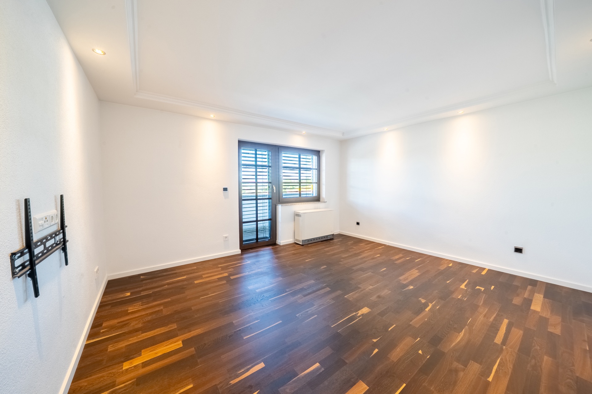 Leeres Wohnzimmer mit dunklem Holzboden, weißen Wänden und Fenster mit Jalousien.