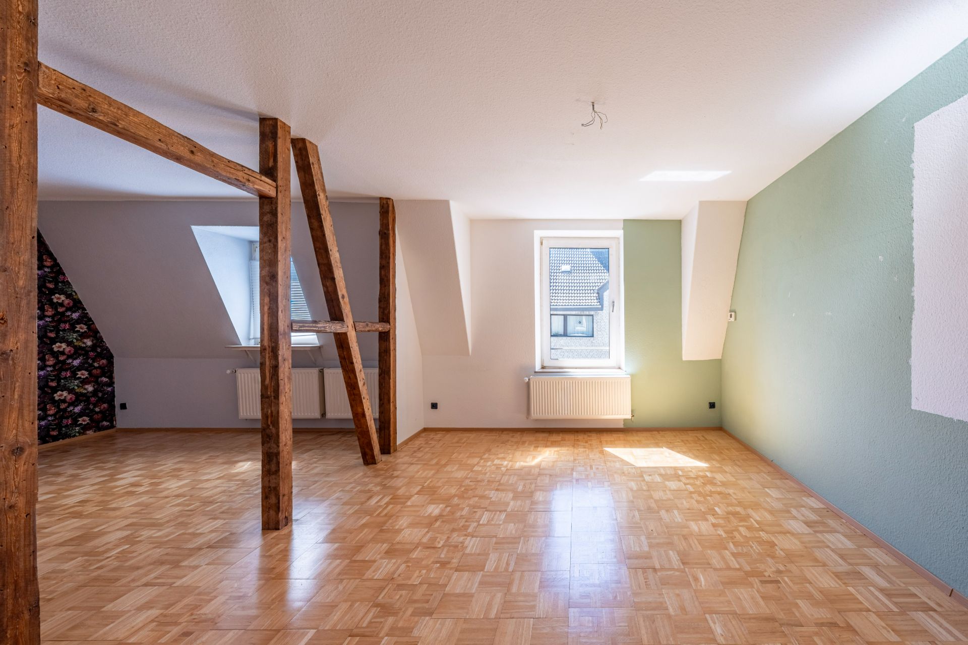 Heller Raum mit Parkettboden, Holzbalken, einem Fenster und schrägen Wänden.
