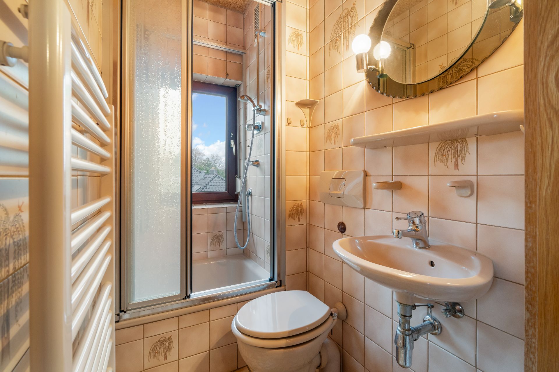 Kleines Badezimmer mit Toilette, Waschbecken, Dusche und Fenster, gefliest in Beige.