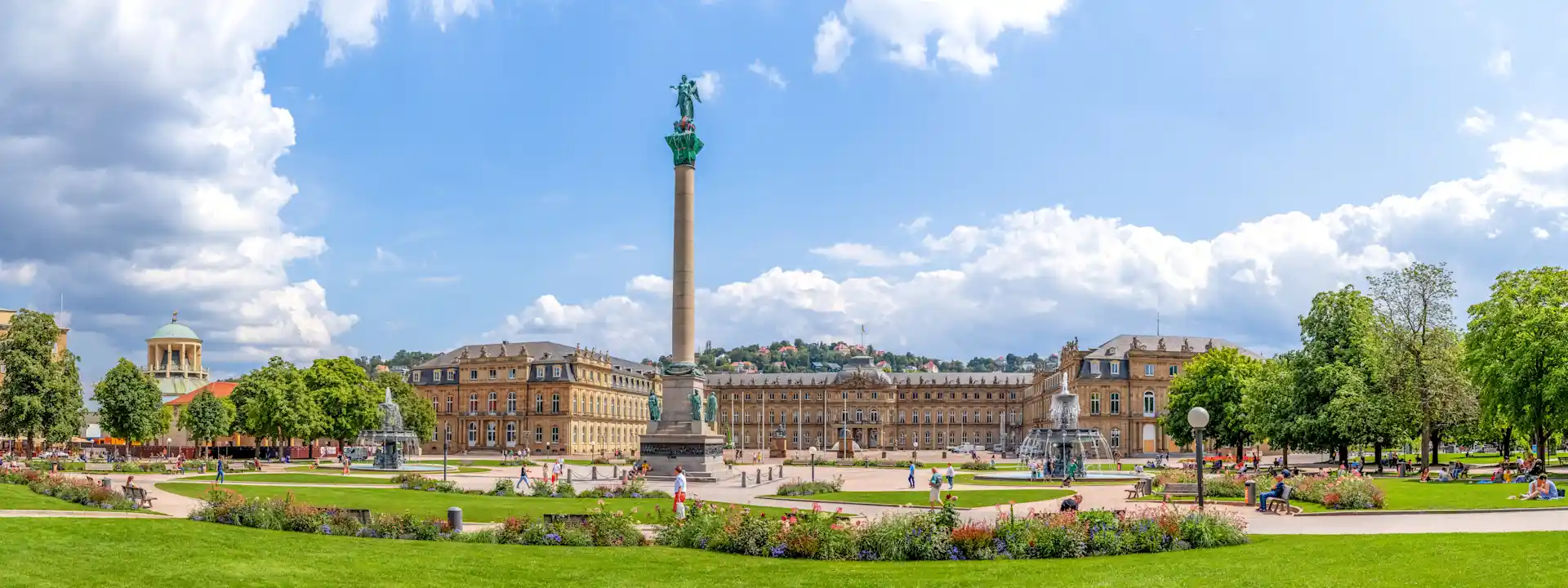 Großer Platz mit Grünflächen, Blumenbeeten, einer hohen Säule und historischen Gebäuden im Hintergrund.