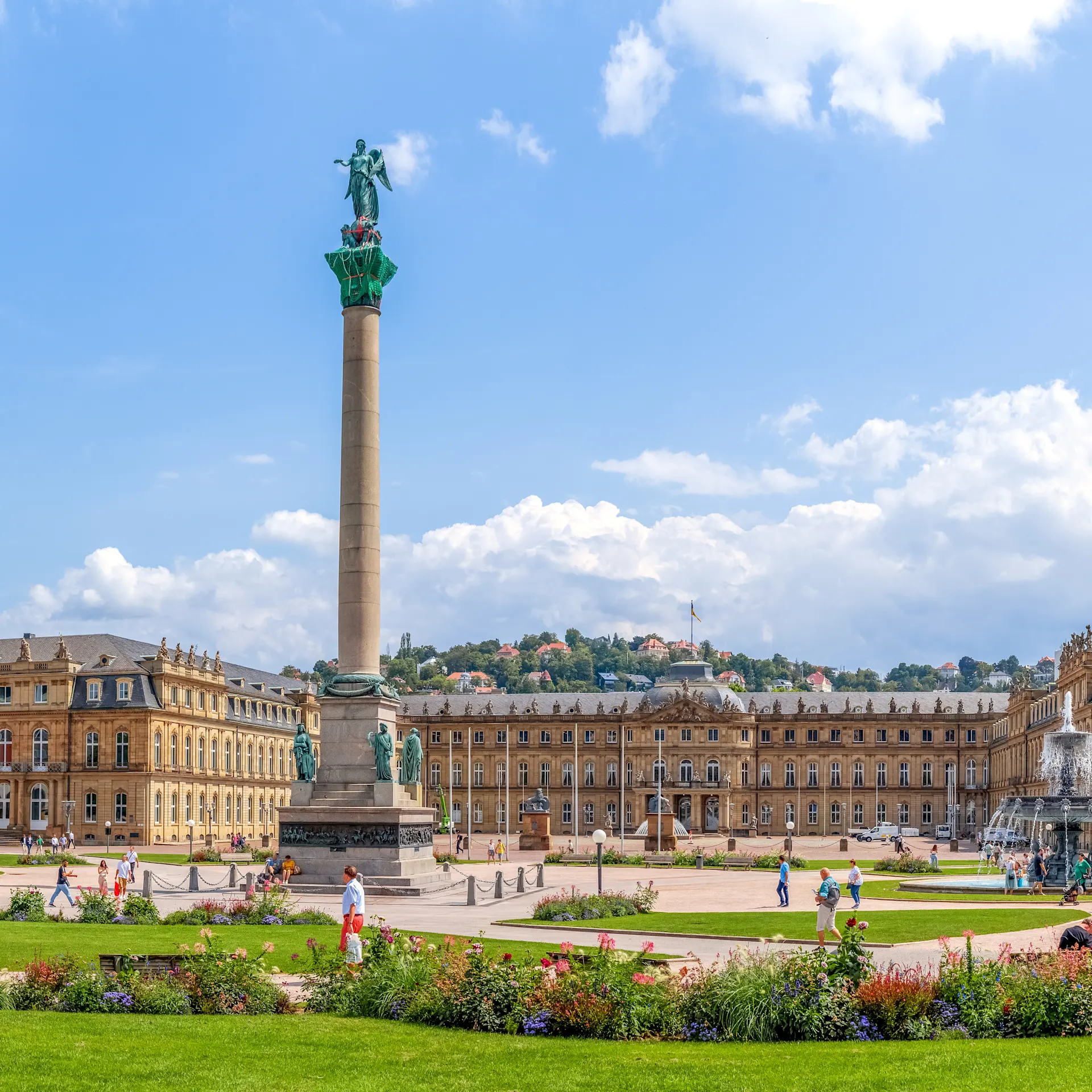 Großer Platz mit Grünflächen, Blumenbeeten, einer hohen Säule und historischen Gebäuden im Hintergrund.