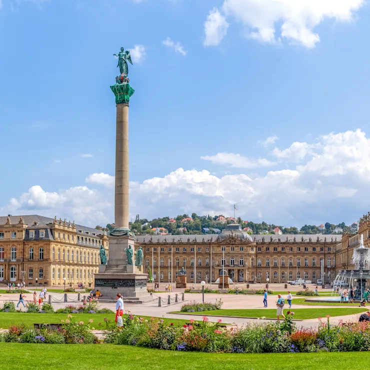 Großer Platz mit Grünflächen, Blumenbeeten, einer hohen Säule und historischen Gebäuden im Hintergrund.