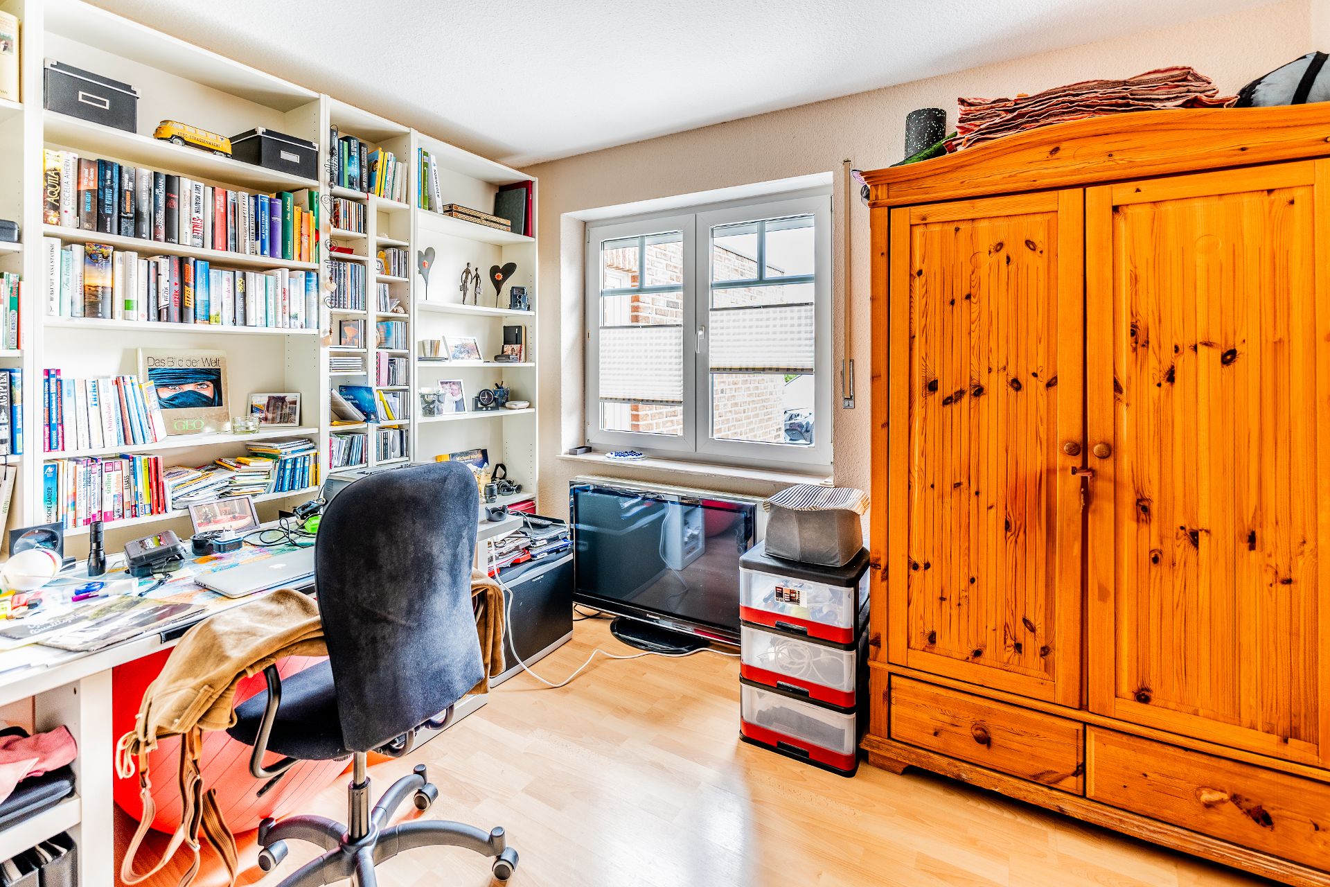 Helles Arbeitszimmer mit Bücherregal, Schreibtisch, Drehstuhl, Fenster und großem Holzschrank.