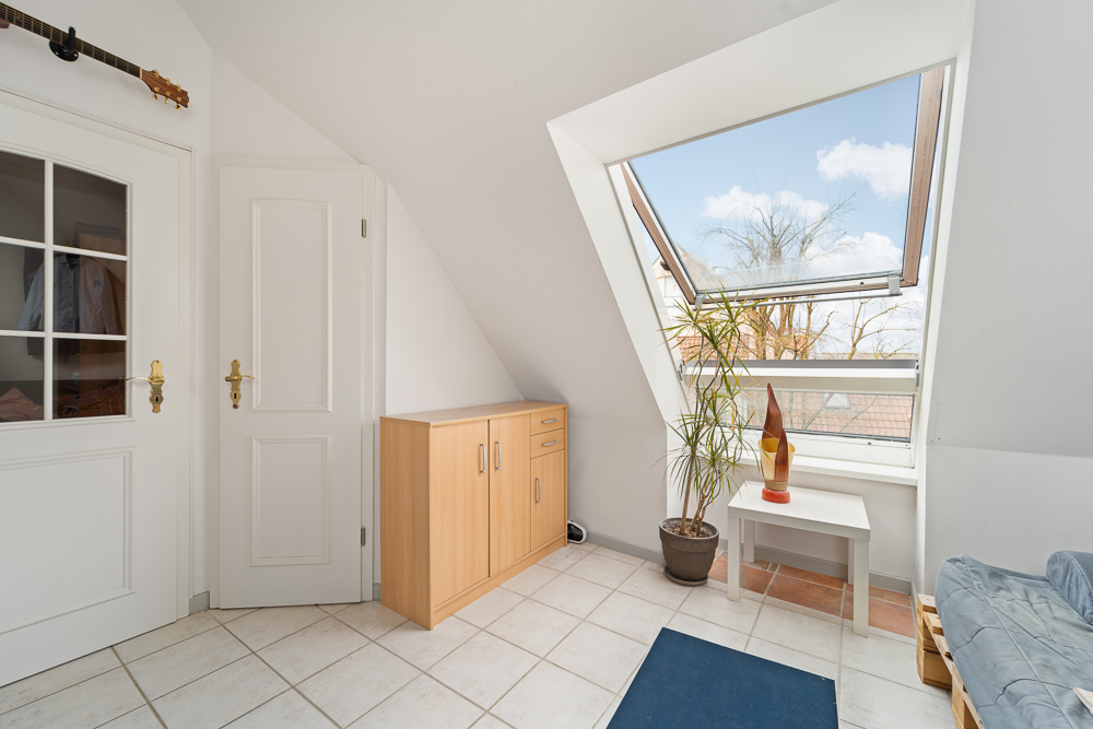 Helles Zimmer mit weißen Türen, Holzkommode, großem Fenster, Pflanze und Tisch mit Deko.