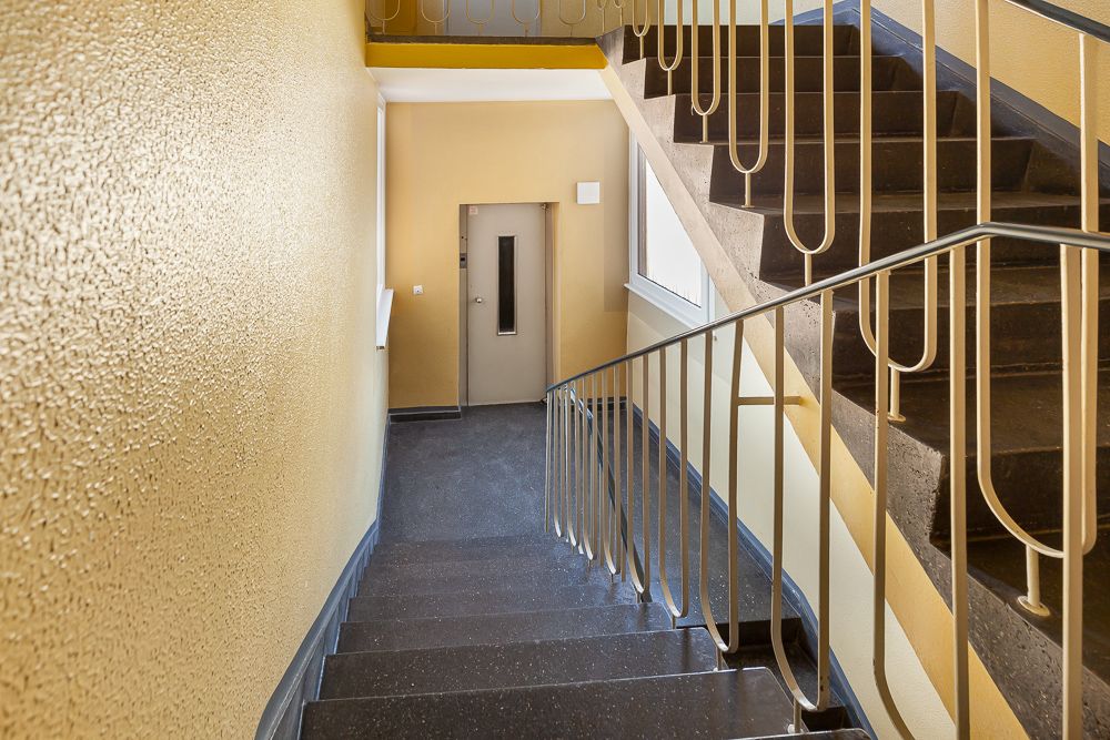 Treppenhaus mit Geländer, gelben Wänden und Aufzugstür im Hintergrund.